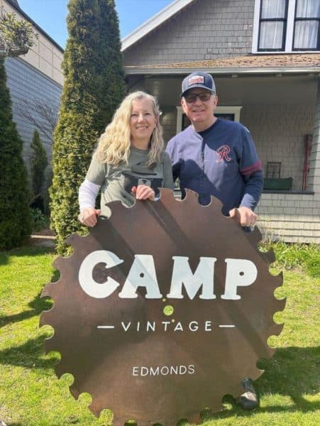 Owners Christine and Logan Harris holding the rusted saw-blade Camp Vintage sign outside the bungalow